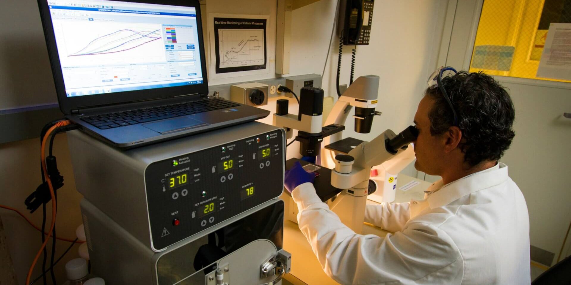 A person looking into a microscope connected to a computer