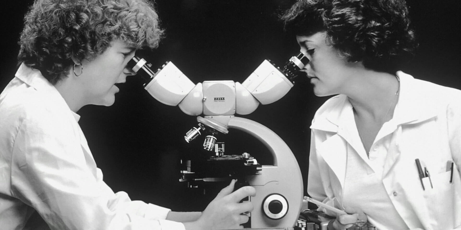 A black and white photo showing two women looking into a microscope