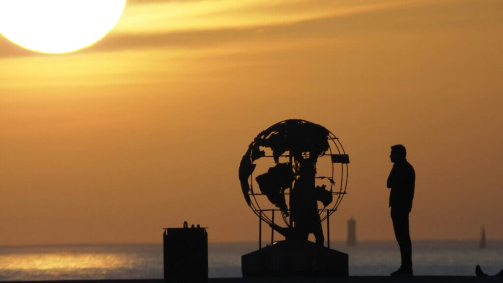 Silhouette of a man and a metal globe by the water with the sun in the background during sunset