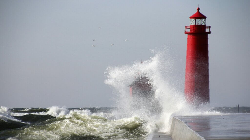 Waves crushing a red light house under grey sky